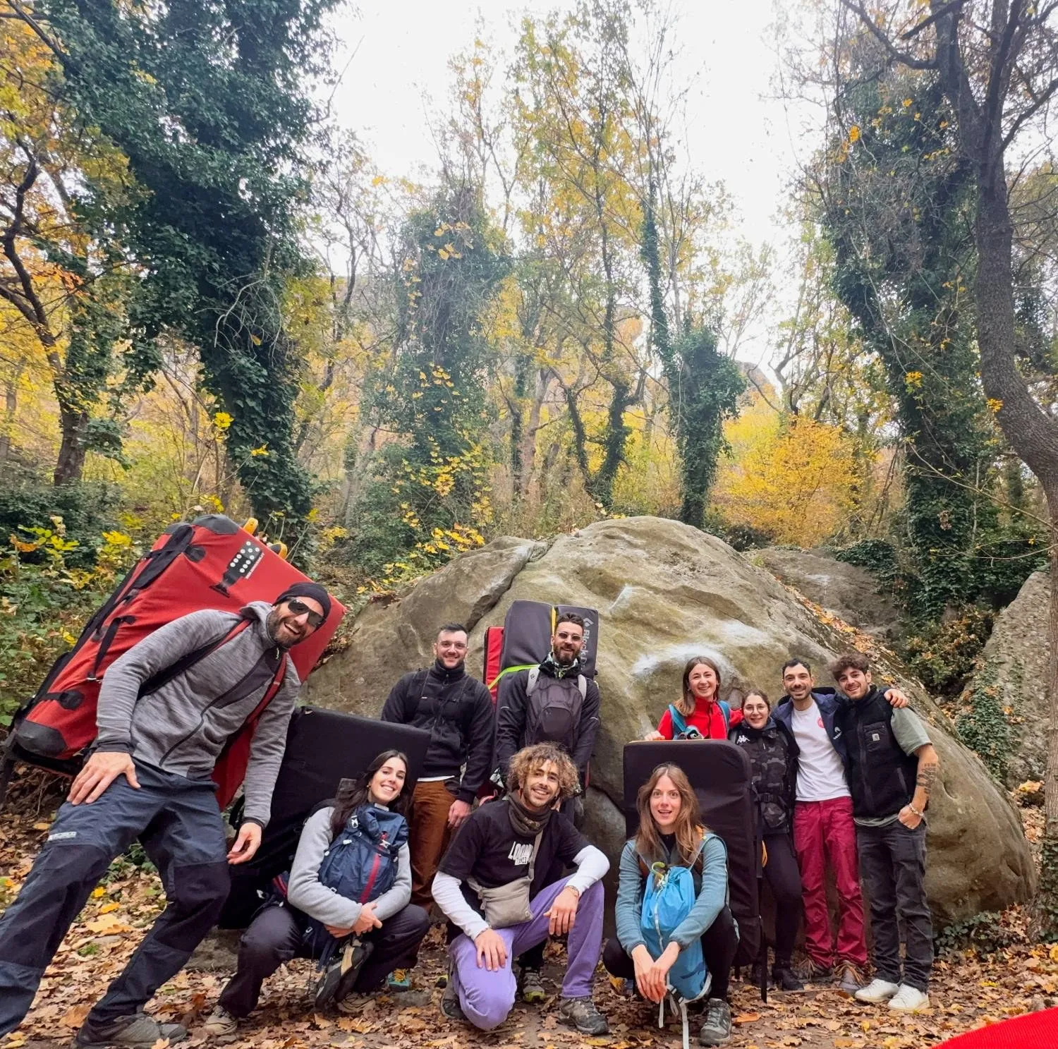 Un gruppo di amici con i crash pad sulle spalle davanti a un masso nel bosco, durante un’uscita di boulder all’aperto