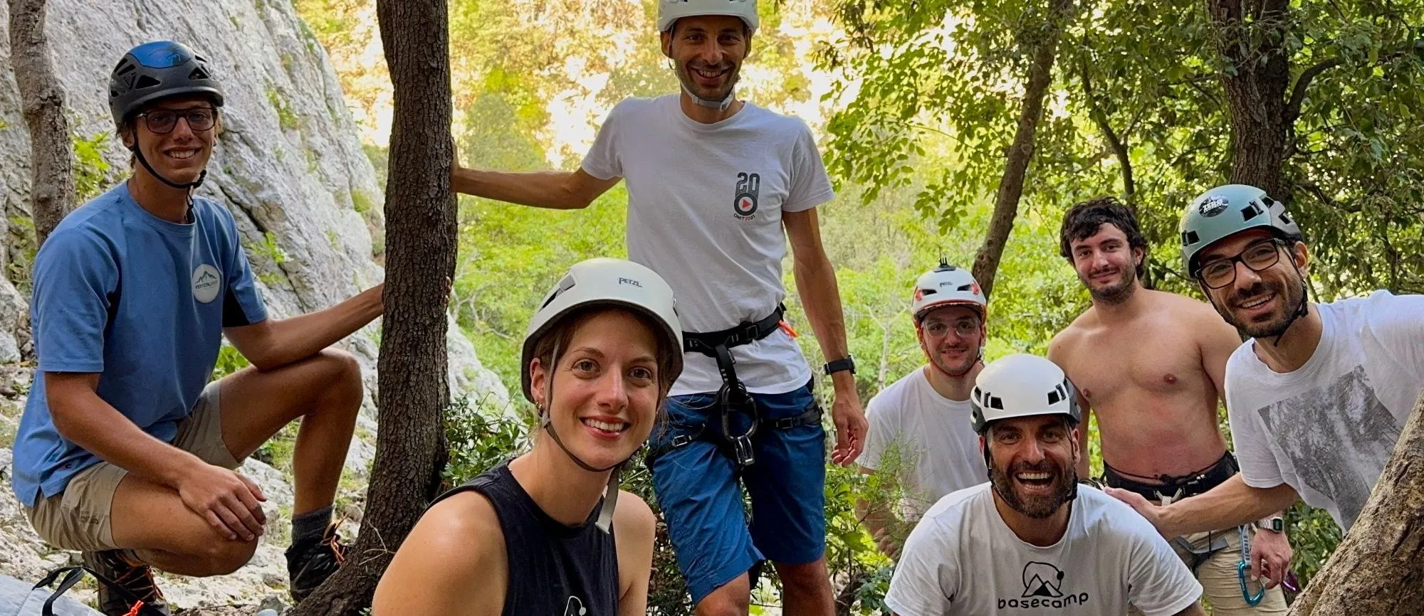 Un gruppo sorridente con caschi e imbraghi nel bosco ai piedi di una falesia, durante un’uscita di arrampicata