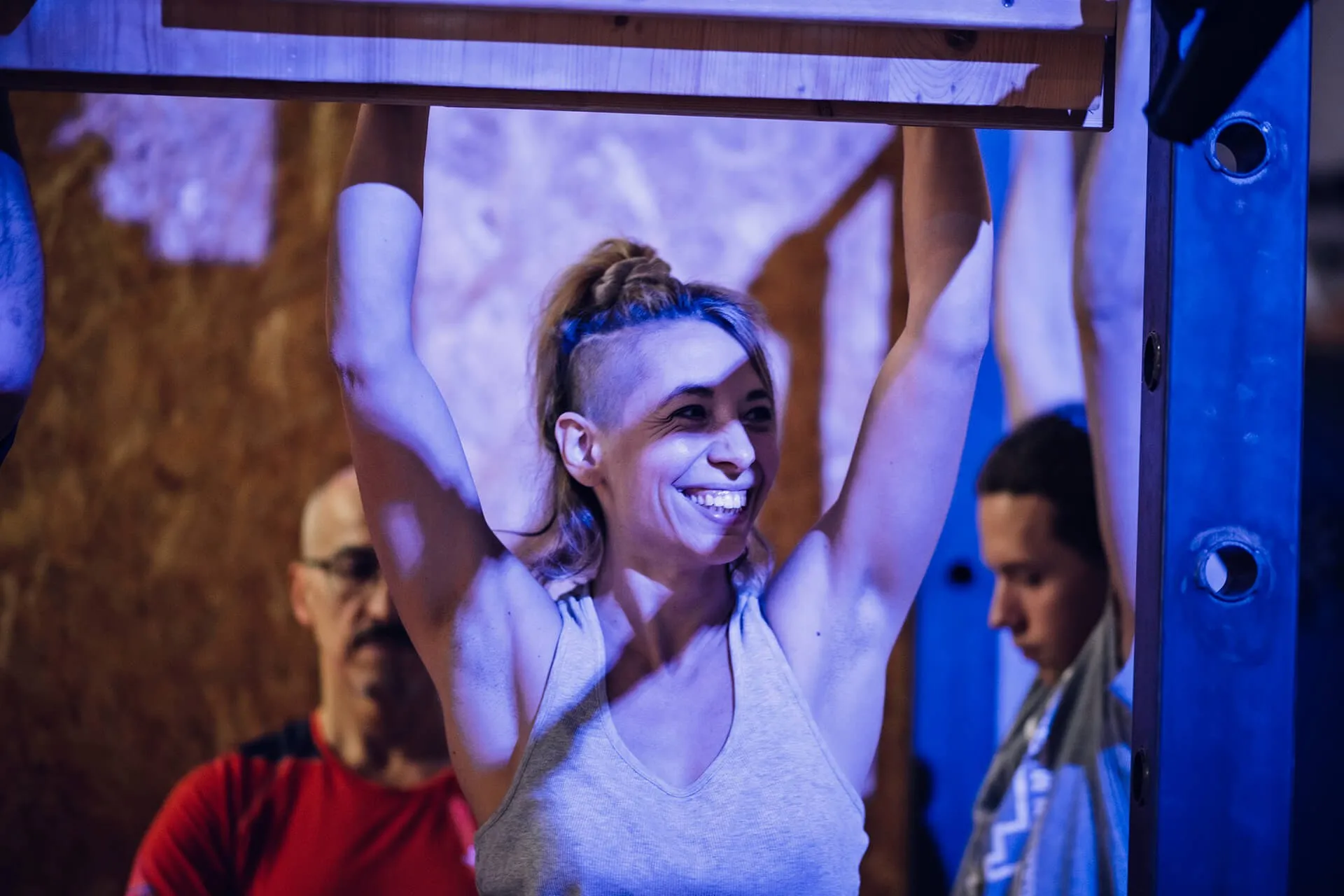 Una ragazza sorridente appesa a una trave di legno durante un allenamento, sotto le luci blu della sala