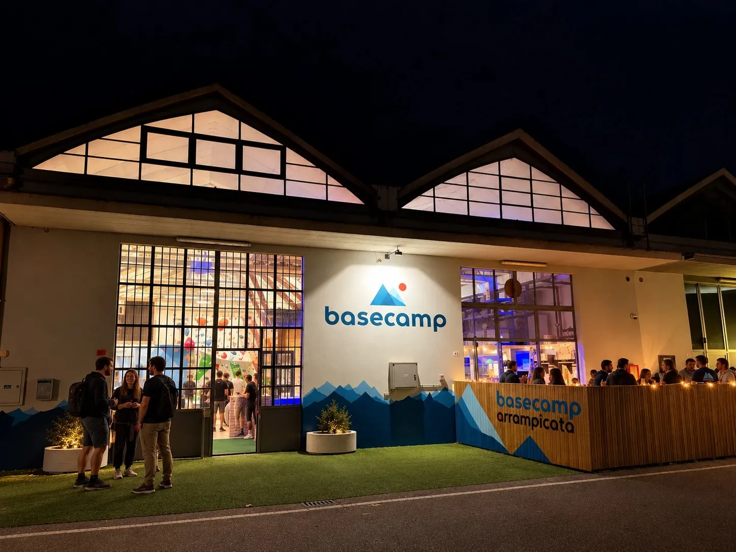 L'ingresso di Base Camp di sera: la facciata con la scritta basecamp e le montagne dipinte, le vetrate illuminate sulle pareti, persone che chiacchierano sul prato davanti e nel dehors