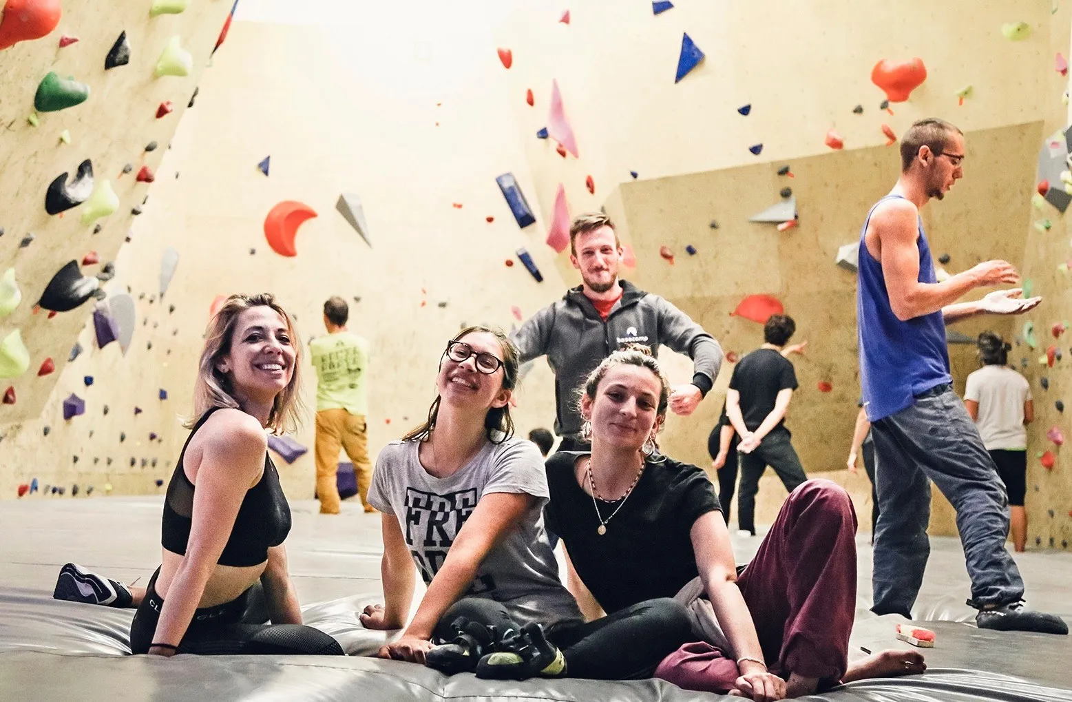 Tre ragazze sorridenti sedute sul materasso e un ragazzo con la felpa basecamp alle loro spalle, mentre in sala altri arrampicano